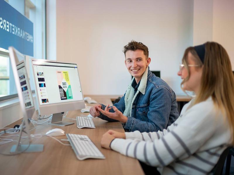 A desk with two people working on computers. One person is talking to the other.