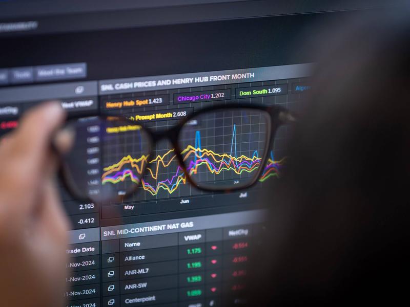 A computer screen displaying stock market data and graphs, viewed through a pair of glasses held in someone's hand.
