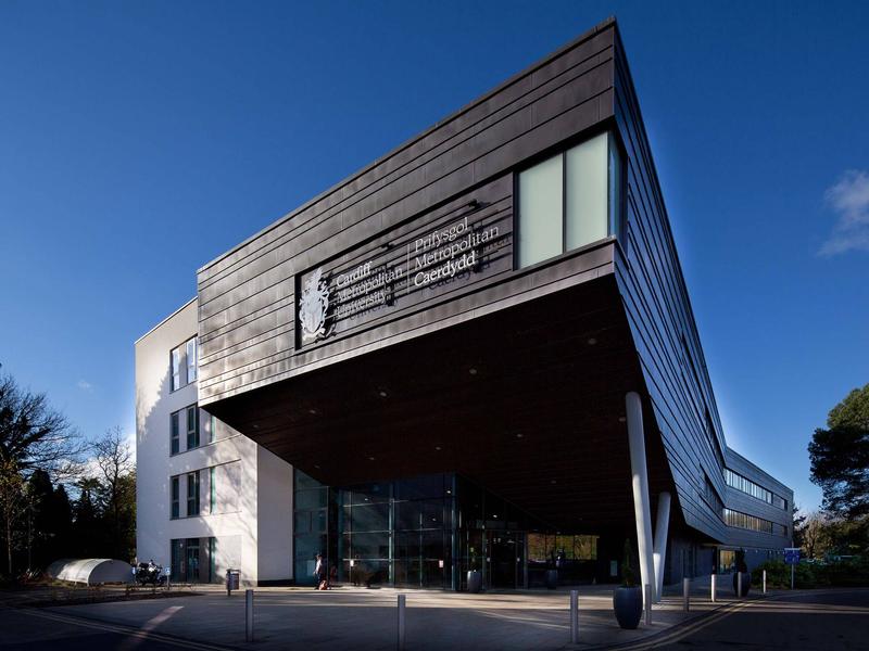 Modern building with angular architecture, featuring a large sign that reads Prifysgol Metropolitan Caerdydd. The structure has a mix of dark panels and white walls, with glass windows. Trees and a blue sky are visible in the background.