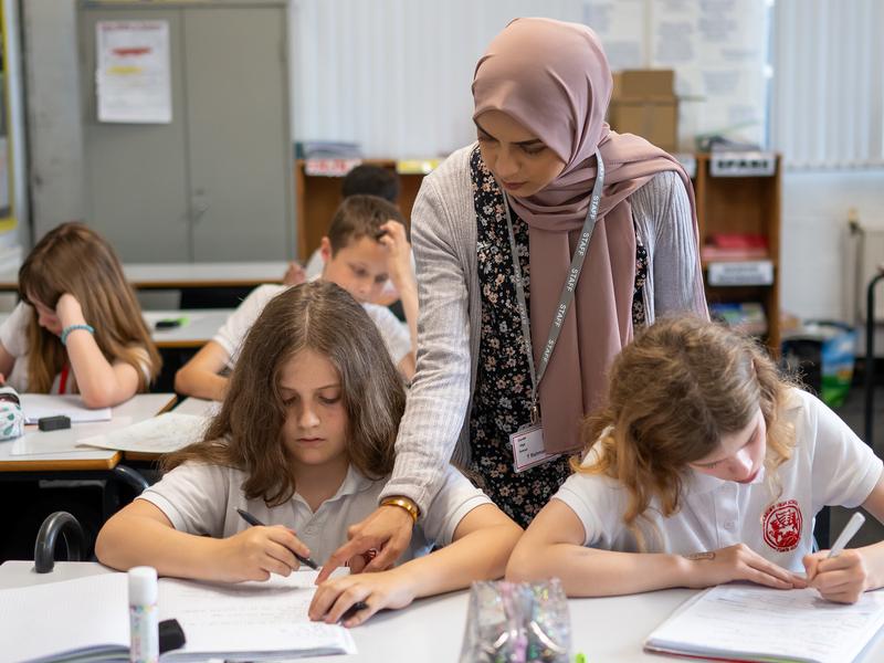 A teacher stands over two pupils writing at their desks.