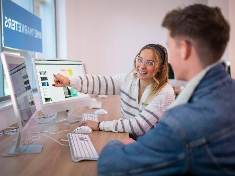 A desk with two people working on computers. One person is pointing to a computer screen while turning to face the other person.