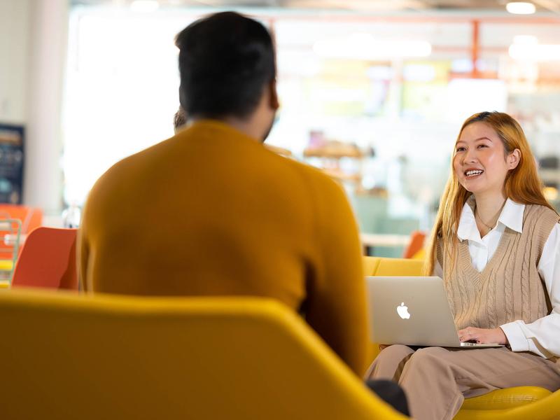 A young woman sits on a yellow chair facing another person. A Mac laptop computer is resting on her lap.