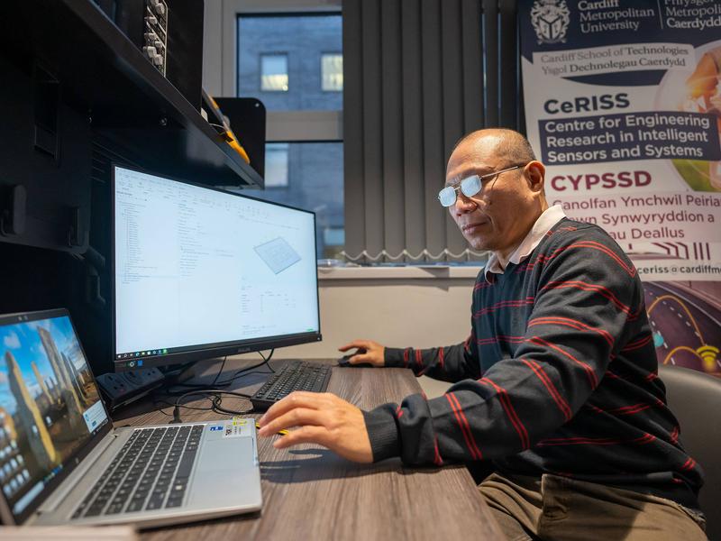 A person sitting at a desk uses a computer workstation. Their right hand controls the mouse, while their left reaches for the trackpad of an open laptop computer.
