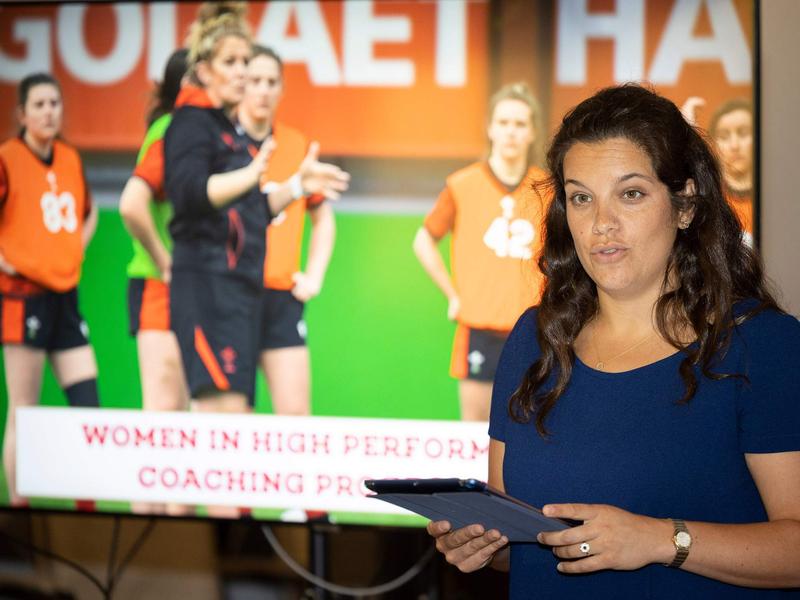 A woman holding an iPad stands in front of large display screen. On the screen are a group of women's rugby players.