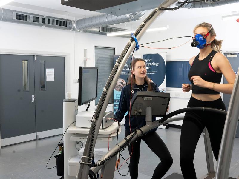 A person runs on a treadmill while wearing a breathing mask. A second person stands alongside, next to a computer display and other equipment.