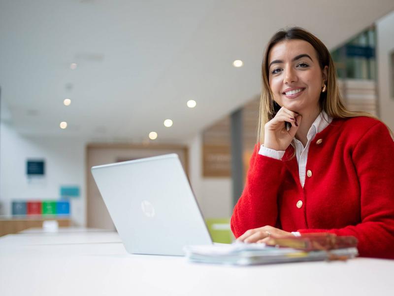 A person is smiling while sitting at a table with a laptop in front of them. They are in a bright, modern setting.