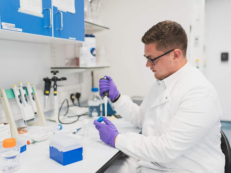 A person in a lab coat uses a pipette to dispense a sample into a container.