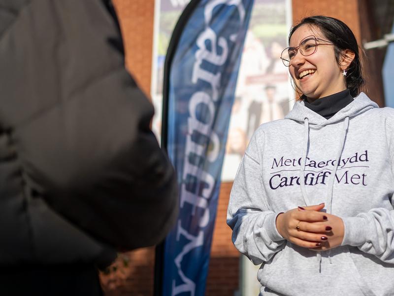 A young woman in a grey Cardiff Met-branded hoodie stands in conversation