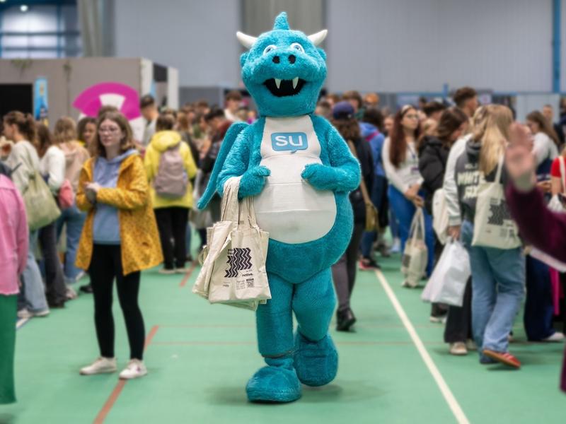 The Students' Union mascot, a light blue and white dragon, holding a tote bag in the middle of a busy Freshers' Fayre