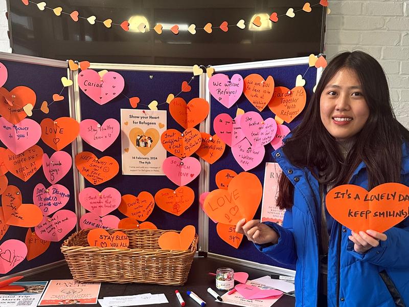 A woman holding a paper heart card stands in front of a display stand filled with paper hearts containing messages of support for refugees