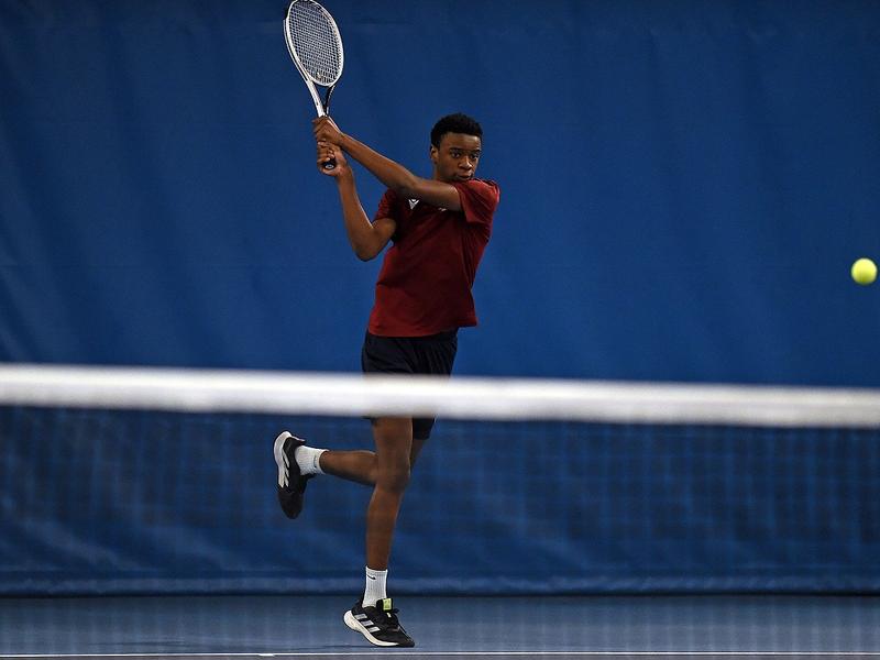 A young man in Cardiff Met exercise t-shirt swings a racket at a tennis ball