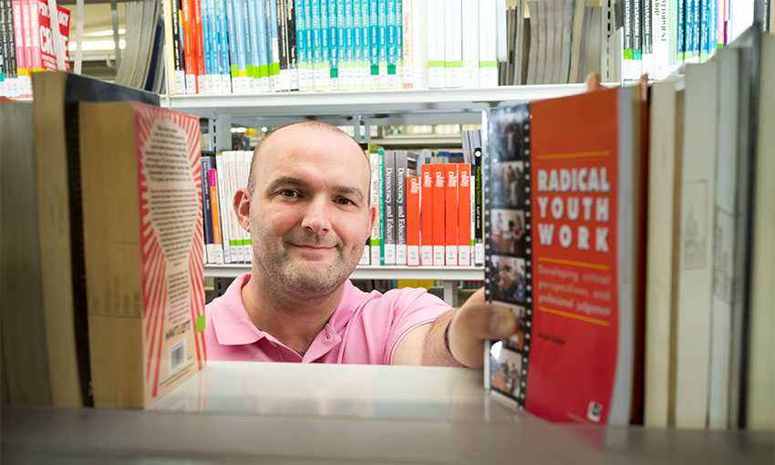 Phil Jones reaches for a book on a library shelf. He is pictured from the other side of the bookcase in a gap between the books