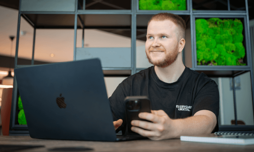 A man wearing a black shirt is seated at a table with a laptop and a cell phone in front of him.