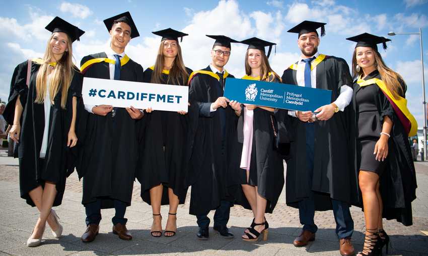 A group of students in graduation caps and gowns stand holding Cardiff Met signs