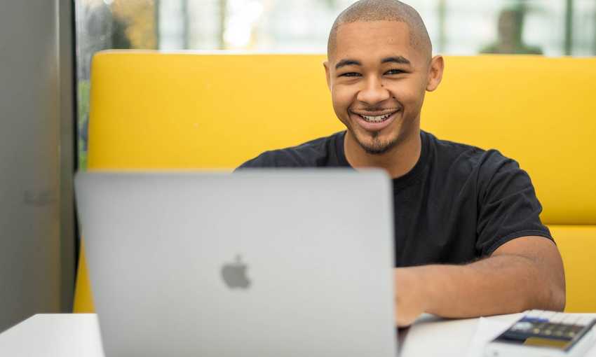 A person is smiling while sitting at a table with a laptop in front of them. They are in a bright, modern setting, with a yellow cushioned seating area behind them.