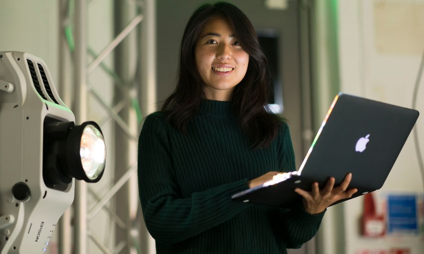 A Woman in a dark green jumper stands holding an open laptop