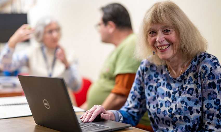An elderly woman works from a laptop in at a busy desk