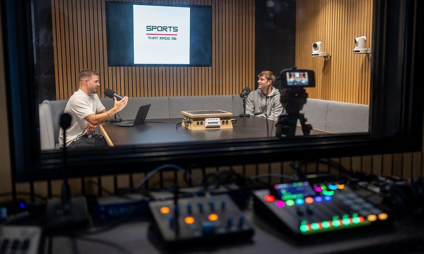 Two young adults sit at far ends of a table talking to one another recording a podcast in a sound booth