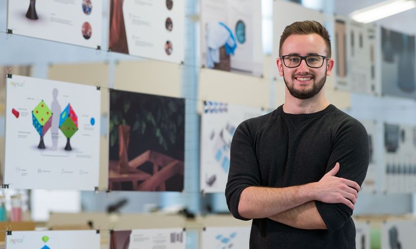 A young adult stands in front of a glass display board featuring pages of designs