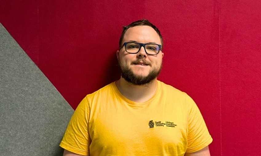 A man in a yellow shirt and glasses poses in front of a red and gray wall.