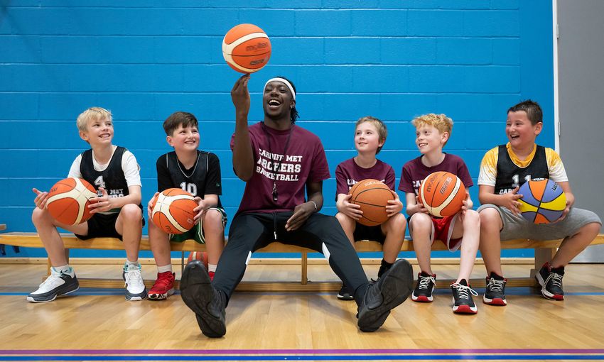 A sports coach spins a basketball on his fingers while four pupils, all holding a basketball on their laps, watch on
