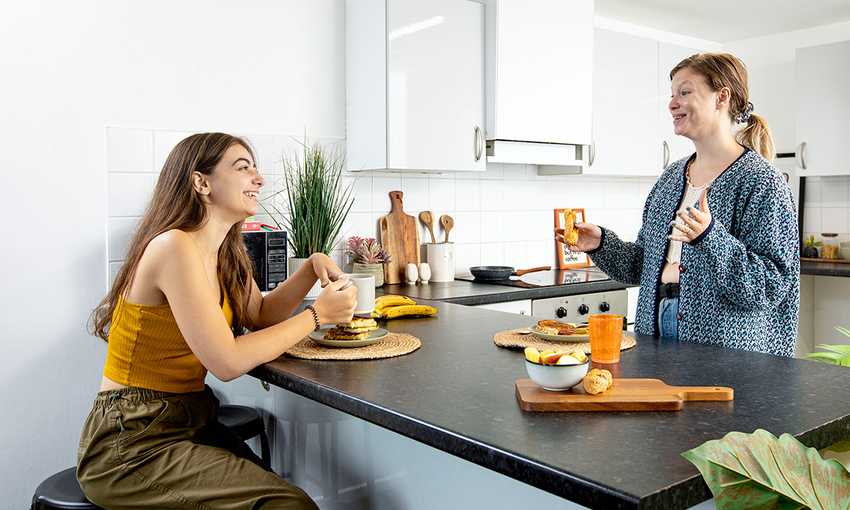 Two female students engaged in conversation while sitting at a kitchen counter, surrounded by kitchen items and warm lighting.