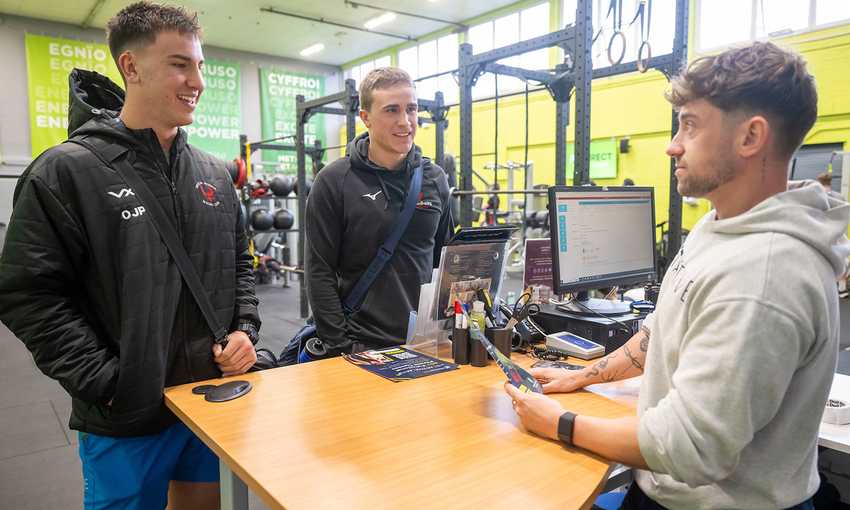 Two men engage in conversation with another man in a gym setting, surrounded by fitness equipment and weights.