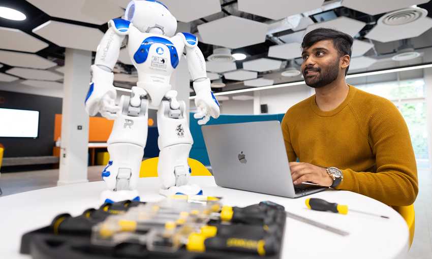 A man at a table using a laptop, accompanied by a robot, engaged in a technological task.