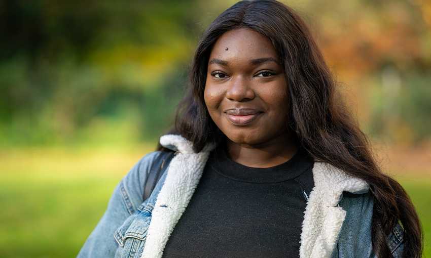A young woman wearing a black shirt and a denim jacket, standing confidently with a casual demeanor.