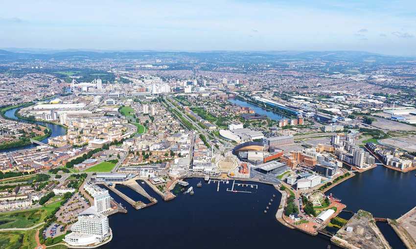 Aerial view of a vibrant waterfront city with winding rivers, numerous buildings, and a marina. The landscape features green spaces and urban areas under a clear blue sky.