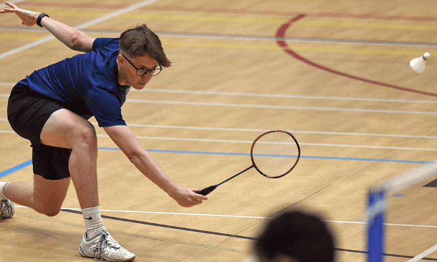 A man wearing a blue shirt and black shorts is actively playing badminton on a court.