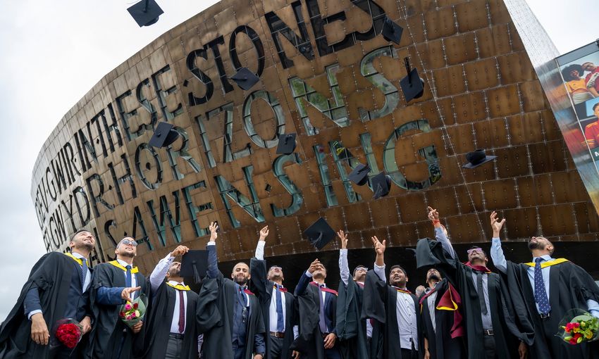 A row of student toss their graduation caps into the air in front of the Wales Millennium Centre sign and entrance