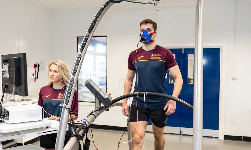A man is walking on a treadmill wearing a blue mask for a fitness test. A woman stands nearby, operating a computer connected to the equipment. Both are in a lab setting wearing maroon and navy sports attire.