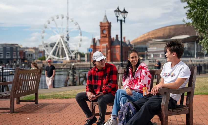 Three students sit on a bench in Cardiff Bay. Behind them is a Ferris wheel, the Pierhead Building and Wales Millennium Centre.