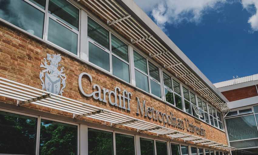A building with large windows featuring the sign Cardiff Metropolitan University on a brick wall. A blue sky with clouds is visible above.