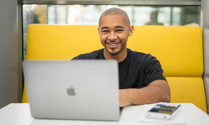 A person is smiling while sitting at a table with a laptop in front of them. They are in a bright, modern setting, with a yellow cushioned seating area behind them.