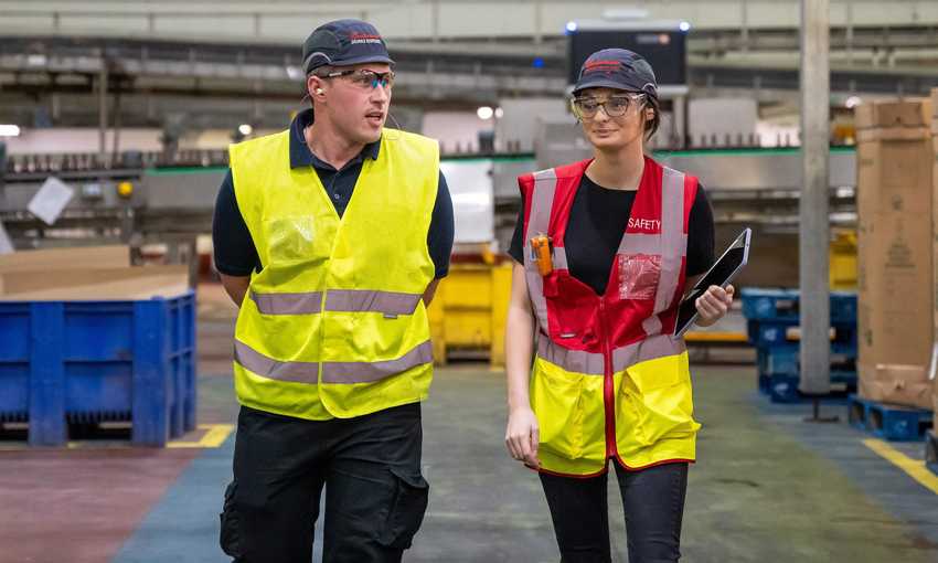 Two people in high vis vests walking on a factory floor. In the background are conveyer belts and cardboard boxes on pallets.