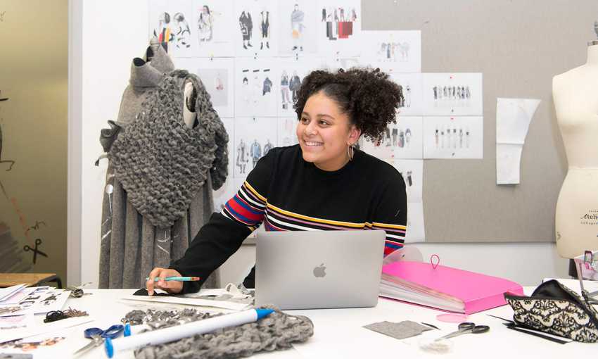 A young woman sits at a desk with a laptop computer and selection of fabric swatches. Behind her is a mannequin and wall of clothing designs.