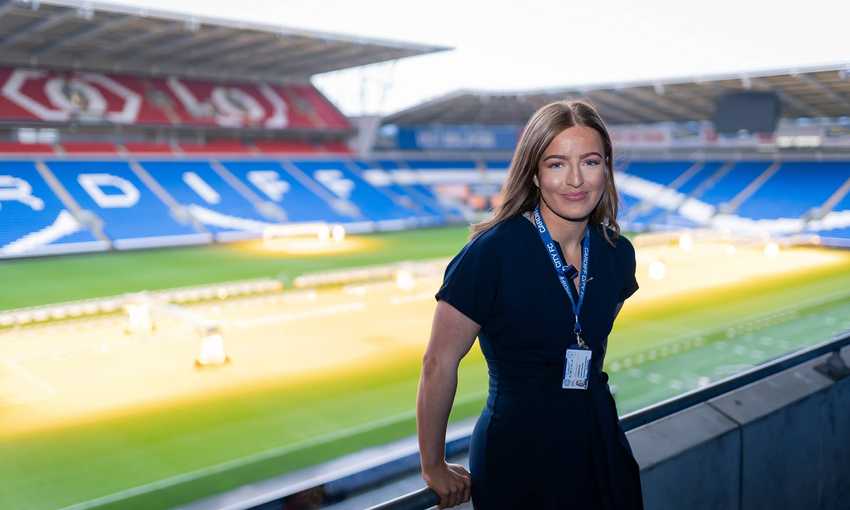 A woman in a stands smiling on the balcony of an empty stadium. The stadium seats are predominantly blue, with some red sections, and green grass covers the field. She wears an ID lanyard around her neck.