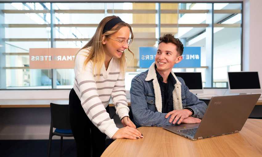 A person sits at a table in front of a laptop computer. Another person stands next to them laughing.