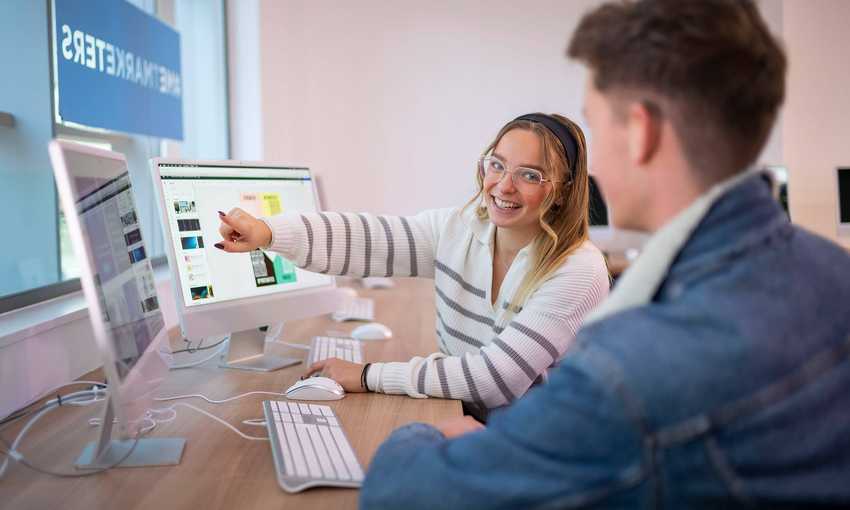 A desk with two people working on computers. One person is pointing to a computer screen while turning to face the other person.