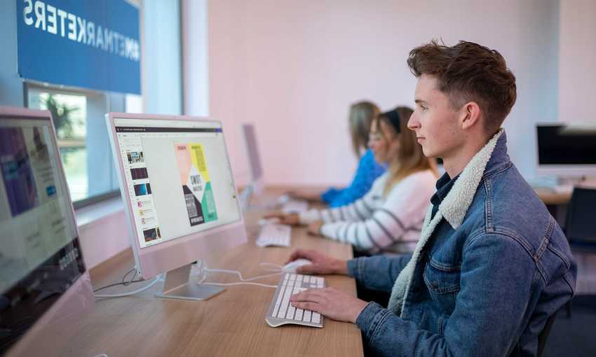A person sat at a desk in front of a Mac computer. In the background are two other people also using computers.