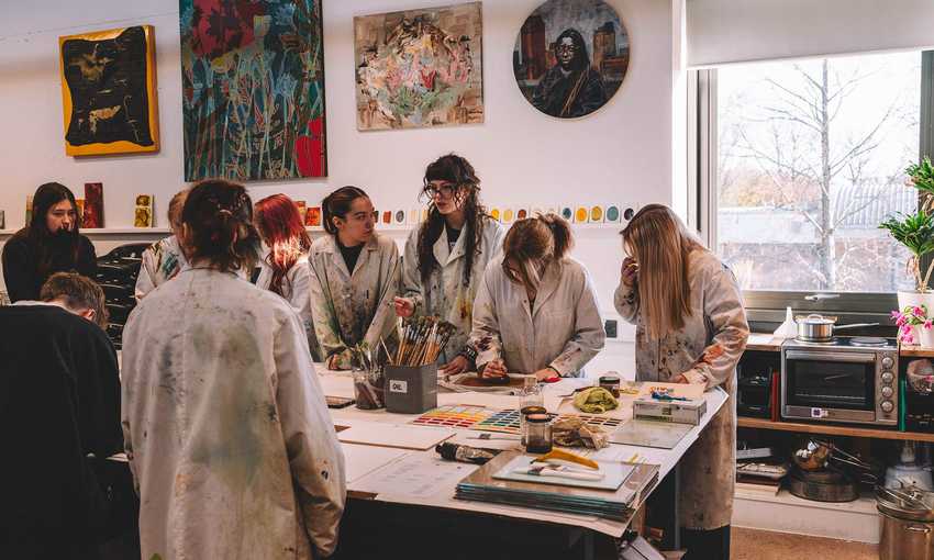 A group of students gather around a large table on which there are brushes and other painting materials. The students are wearing white coats spattered with paint.