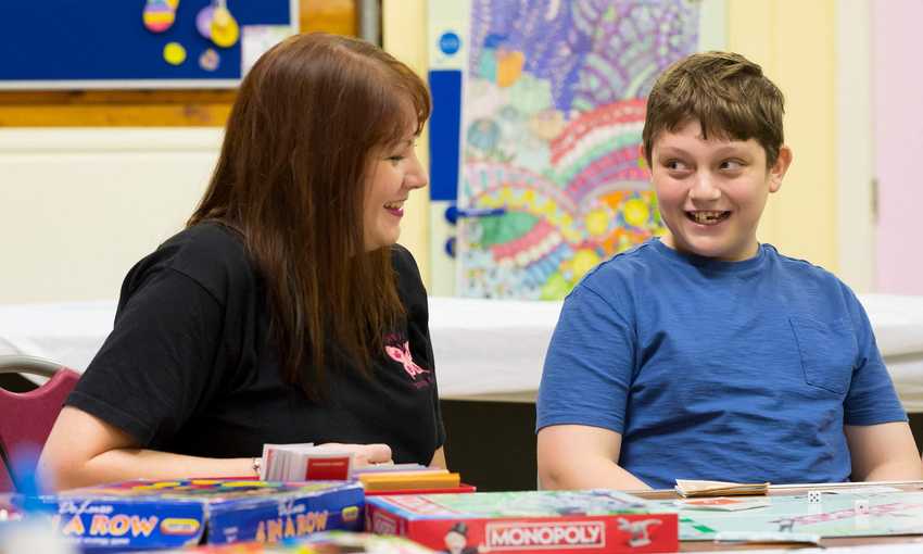 A woman sits next to a child. On the table in front of them is a selection of board games.