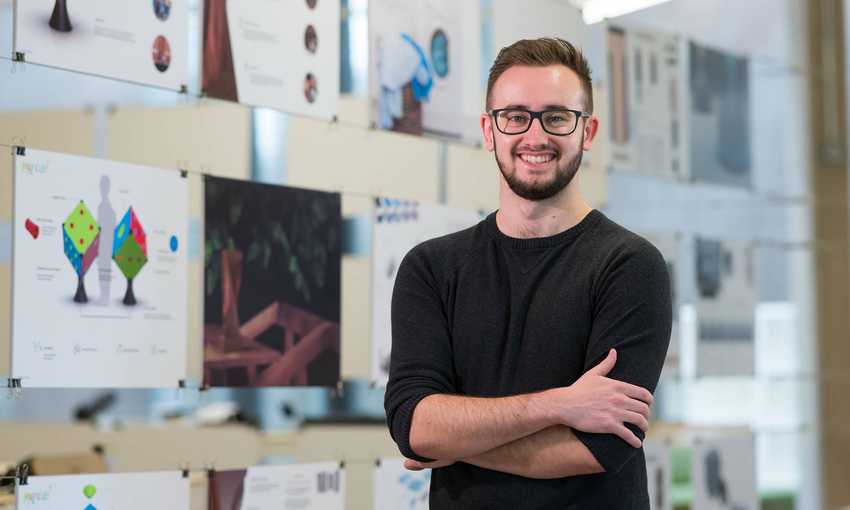 A young man in a dark sweater stands with his arms folded. Behind him is a display of drawings and designs.