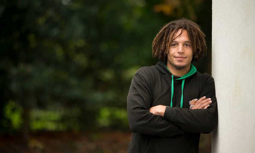 A young man wearing a dark grey hoodie leans against a wall with his arms folded.