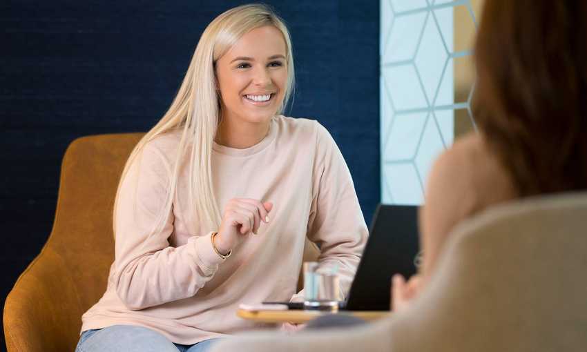 A young woman with long blonde hair sits on a chair talking to another person. In front of her is a table with an open laptop computer and a glass of water.