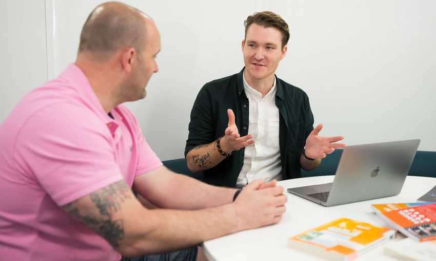 A young man sits at a round table talking to another man sat next to him. On the table is an open laptop computer and several textbooks.