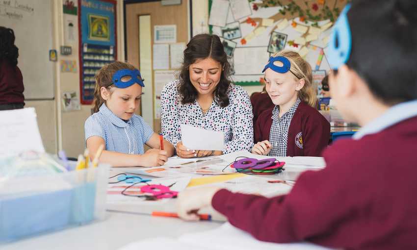 A teacher between two pupils sitting at a desk. The teacher is holding a sheet of paper.