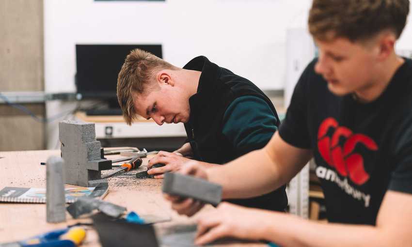 Two students sit alongside each other at a table. They are filing and shaping grey blocks of material.